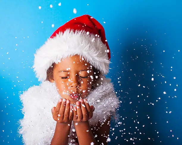 Photo of African American little girl in Santa Claus hat blowing snowflakes