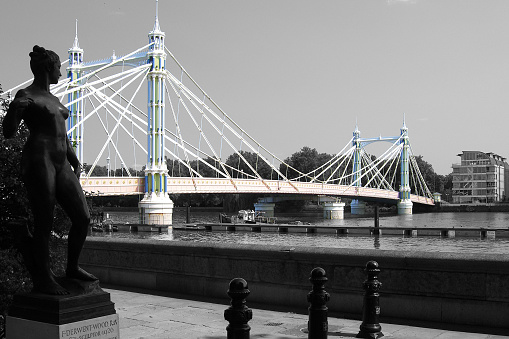 The Albert Bridge designed and built by Rowland Mason Ordish in 1873, connecting Chelsea on the north bank of the Thames river to Battersea on the south bank. On the left side, the statue of the Atalanta (credited as “Chelsea Embankment statue”) by the famous british sculptor Francis Derwent Wood (1871–1926). The whole picture is in black and white except for the bridge.