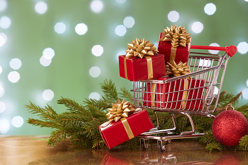 Shopping trolley with gift boxes and brenches of fir-tree with decoration on a green background with light of garland. Christmas and New Year sale.