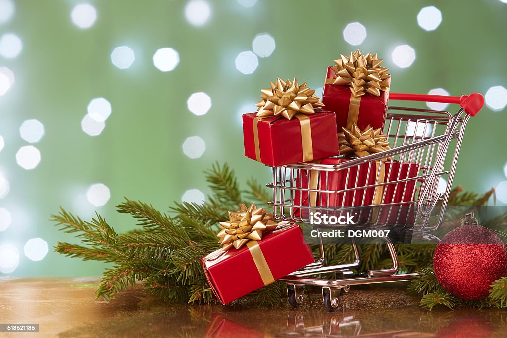 Christmas and New Year shopping. Shopping trolley with gift boxes and brenches of fir-tree with decoration on a green background with light of garland. Christmas and New Year sale. Christmas Stock Photo