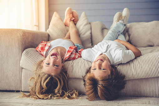 Pretty little girl and boy are lying on their backs on sofa, looking at camera and smiling while playing at home