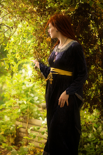 Vertical outdoor shot in autumn garden, of mid adult woman. She has long red hair in dreadlocks and is wearing a blue gown of medieval look. Standing in dark part of garden, pulling back vines to look into brighter area. Concepts of wonder and exploring