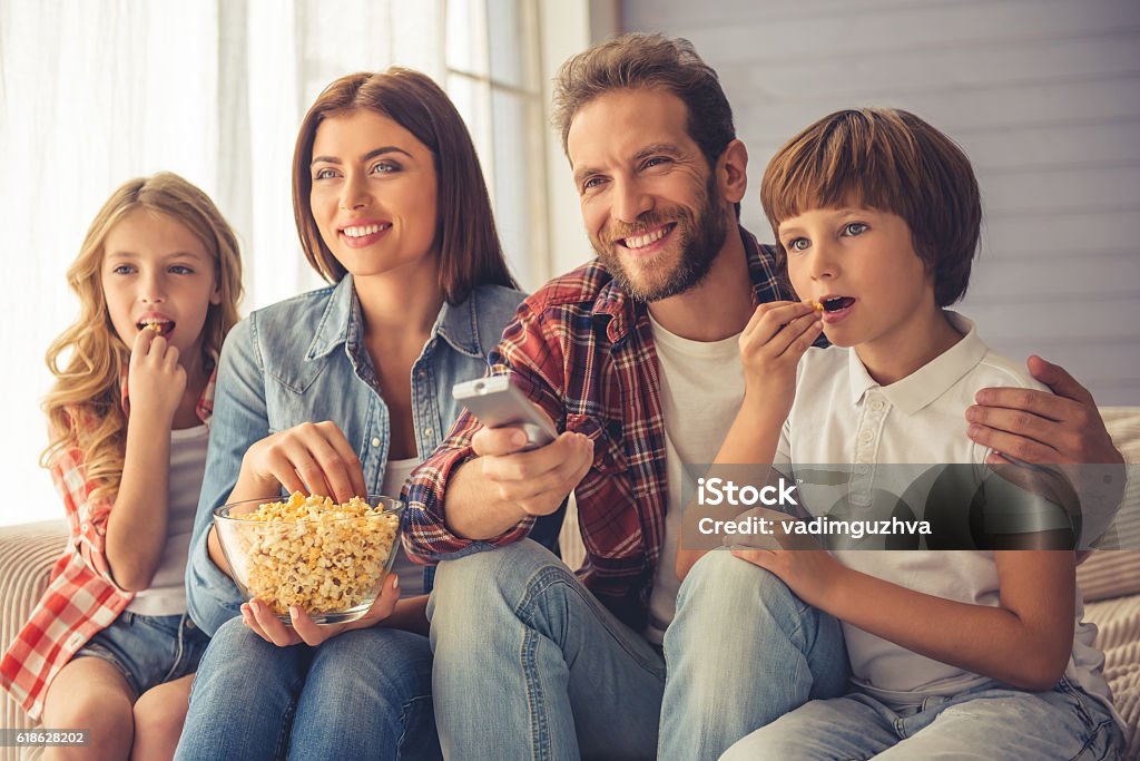 Happy family together Beautiful young parents and their children are watching TV, eating popcorn and smiling while sitting on couch at home Family Stock Photo