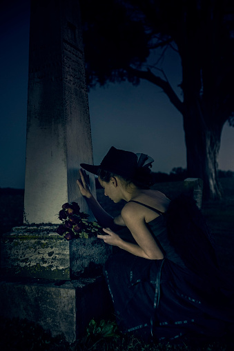 Female in 20s dressed in a long vintage black dress holds a lit lantern as she walks through a tiny cemetery with ancient graves in a corn field in rural Indiana at night in an eerie blue light