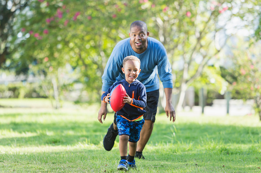 A cute little 3 year old African American boy playing with his father in the park. He is running, carrying an American football, his father chasing him.