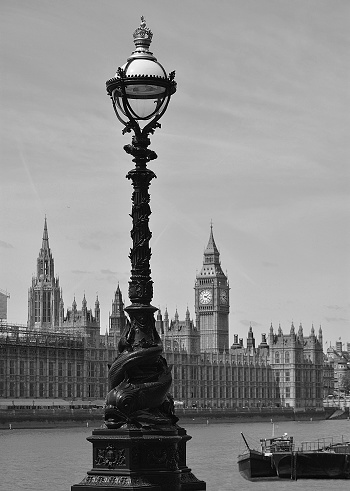 Quaint image in black and white of Westminster Houses of Parliament, Big Ben and the river Thames as the background to one of many ornate street lamps in this are of London, UK. 