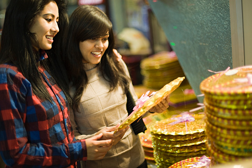 Image of two late teen Asian happy girls of different ethnicity selecting and buying gifts at gift shop just before new year and Diwali festival. Two people, waist up, horizontal composition and selective focus with copy space.