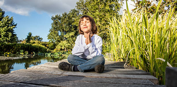 zen young yoga child meditating alone to breathe near water zen young yoga child cross-legged enjoying meditating alone, closing eyes for relaxation and breathing on a wooden bridge near water with blue sky, low angle view mindfulness kids stock pictures, royalty-free photos & images