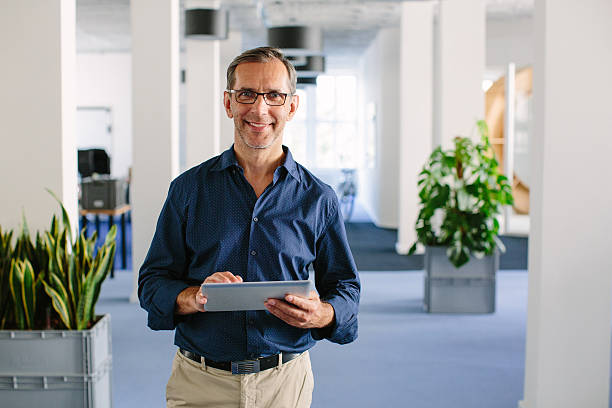 Successful senior businessman standing in office Shot of a successful senior businessman standing in office with digital tablet business casual stock pictures, royalty-free photos & images