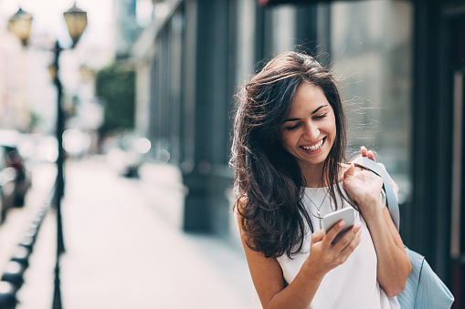 Smiling young woman walking outdoors at urban setting and checking messages.