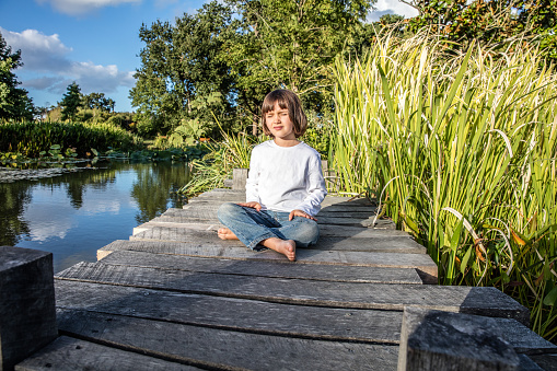 relaxed beautiful young 5-year old child cross-legged with bare feet breathing alone, closing eyes for yoga and meditation near water and trees