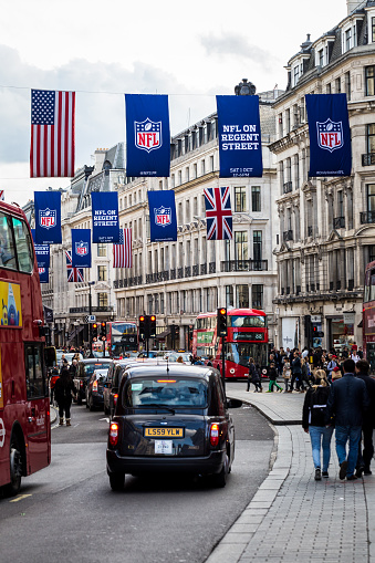 London, UK - October 15, 2016: Lots of iconic red London buses and black cabs on the road in Regent Street, one of the most famous shopping streets in London. The streets are completely thronged with people, while above them the union jack flag of the UK, the American flag, and a banner with the NFL - National Football League - logo, recede into the distance.