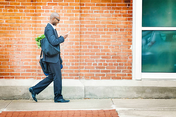 Mature businessman walking back home with groceries Mature businessman walking back home with groceries while social networking on his phone. Full length profile shot in front of brick wall. wall sidewalk city walking stock pictures, royalty-free photos & images