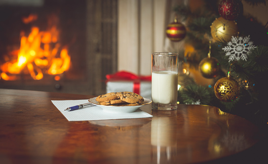 Closeup image of wish list and treats for Santa Claus on table next to burning fireplace