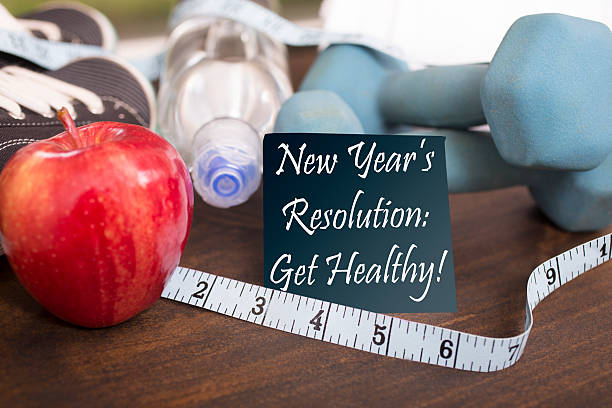来年の1月に健康になる新年の決意。 画像機能:ダンベル、スポーツシューズ、水筒、リンゴ、タオル、木製テーブルの上のテープメジャー。 「新年の決意:健康になる!」を前面に読む粘着音。 フィットネスコンセプト。