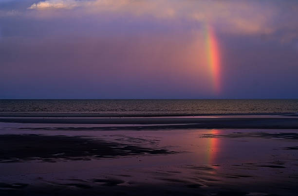 Clearing thunderstorm moving eastwards into the Pacific Ocean over Moreton Island is followed by a rainbow reflected in the tidal flats of low tide over Moreton Bay, producing intense colors due to the setting sun in the west. Photographed from Beachmere on the southern end of the Sunshine Coast in Queensland, Australia.