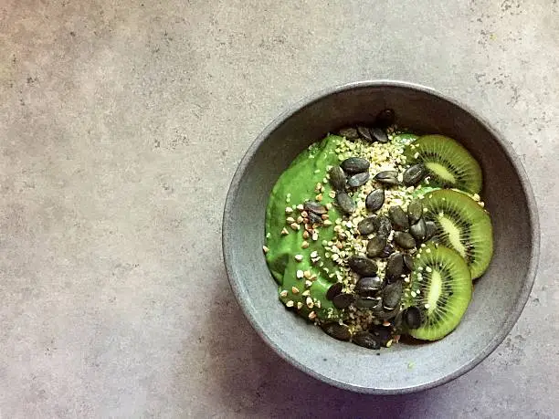A healthy breakfast: a green smoothie bowl in a grey stoneware bowl topped with kiwifruit slices, pumpkinseeds, hempseeds and buckwheat. Smoothie contains spirulina, avocado, banana, kale.