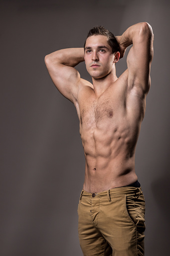 young bodybuilder posing strong muscular torso, chest, abs, arms up. pants, studio.