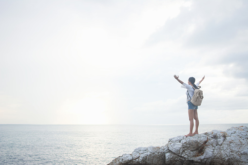 A young Asian woman hiker on vacation stands by the beach wearing a backpack on a coastal rock at sunrise with her arms stretched looking toward the rising sun. Travel, freedom and new beginnings.