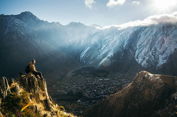 Backpacker sit on cliff edge and looks at mount valley Young tourist in bright hat, black trousers with a backpack sit on cliff's edge and looking at the misty mountain village and glacier at sunrise, Stepantsminda, Georgia georgia landscape stock pictures, royalty-free photos & images