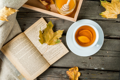 Cozy autumnal mood, warm autumn. A cup of hot tea with lemon and ginger on a rustic table, plaid, yellow leaves, honey and a book to read. Top view 