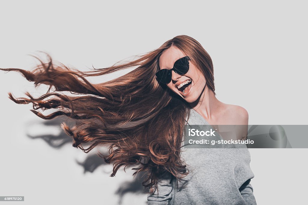 Hair like fire. Attractive young smiling woman in sunglasses and with tousled hair looking away while standing against grey background Long Hair Stock Photo