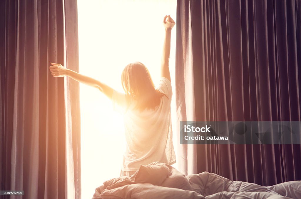 Woman stretching near bed after wake up Woman standing near the window while stretching near bed after waking up with sunrise at morning, back view Waking up Stock Photo