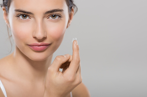 Young woman holding contact lens on index finger with copy space. Close up face of healthy beautiful woman about to wear contact lens. Eyesight and ophthalmology concept.