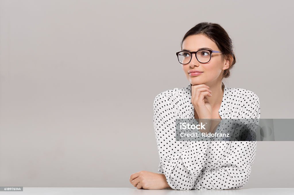 Young woman thinking Beautiful young businesswoman wearing glasses and thinking with hand on chin. Smiling pensive woman with eyeglasses looking away isolated on grey background. Fashion and contemplative girl smiling and meditating on project. Women Stock Photo