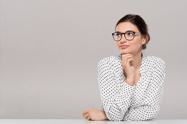 Young woman thinking Beautiful young businesswoman wearing glasses and thinking with hand on chin. Smiling pensive woman with eyeglasses looking away isolated on grey background. Fashion and contemplative girl smiling and meditating on project. smirking stock pictures, royalty-free photos & images