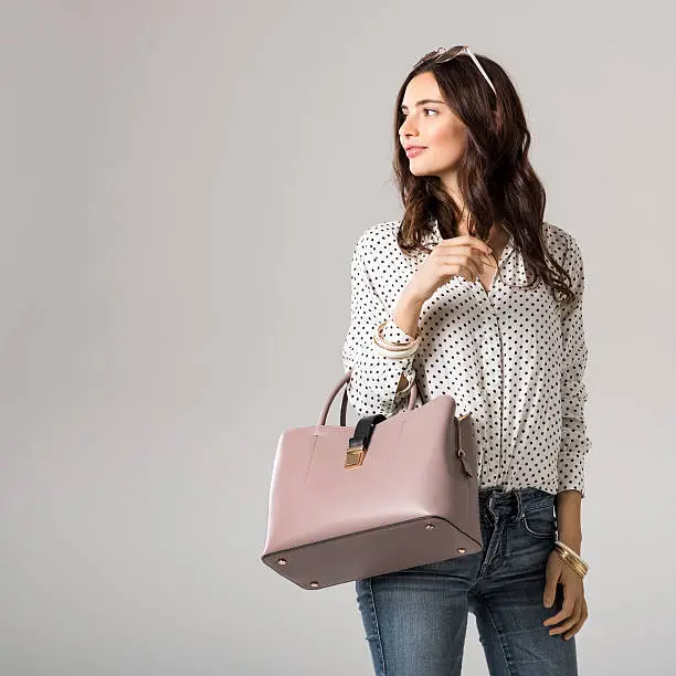 Young glamour woman wearing polka dot shirt and jeans posing with pink handbag. Beautiful stylish girl holding bag and looking away with copy space. Fashion woman holding peach bag with sunglasses on head.