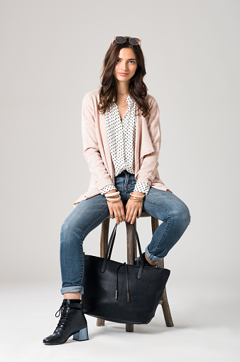 Portrait of young brunette woman sitting on wooden stool and looking at camera. Smiling fashion girl wearing stylish clothes. Happy beautiful woman with sunglasses and shopping bag sitting on stool.