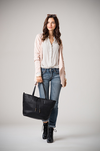 Beautiful cool woman holding black shopping bag and walking on grey background. Brunette fashion girl wearing pink cardigan, jeans and polka dot shirt. Stylish casual woman holding a big bag and looking away.