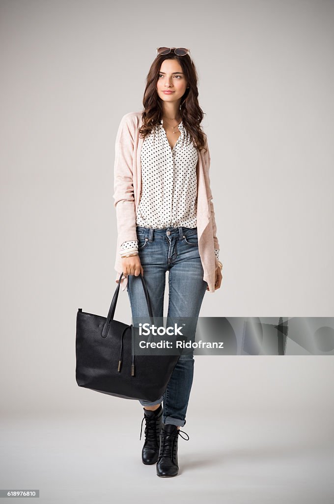 Glamour woman walking Beautiful cool woman holding black shopping bag and walking on grey background. Brunette fashion girl wearing pink cardigan, jeans and polka dot shirt. Stylish casual woman holding a big bag and looking away. Women Stock Photo