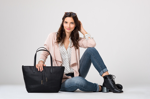 Cool young woman sitting down isolated on grey background. Young fashion woman looking at camera with trendy shopping bag. Stylish girl in casual clothes with accessory smiling.