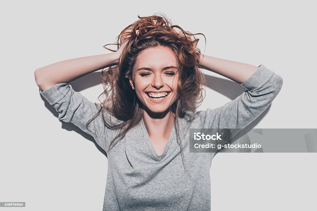 True happiness. Attractive young smiling woman holding hands in hair and keeping eyes closed while standing against grey background Women Stock Photo