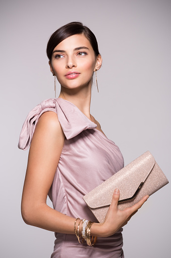 Young beautiful woman in elegant pink dress holding clutch and looking away. Fashion chic woman with purse ready for party. Portrait of elegant girl with luxury accessory isolated on background