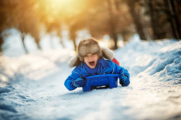 Little boy in funny fake fur hat sliding fast on his sled. The boy aged 6 is screaming with joy. Sunny winter day evening.