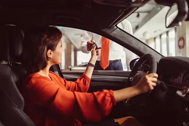 Young woman sitting in new car and taking the keys from the car dealer