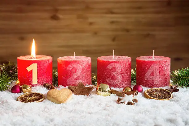 Four numbered red advent candles with first candle lit and christmas decoration lying in snow as template