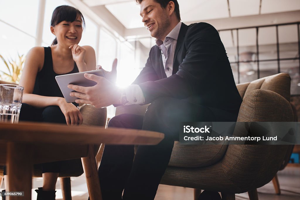 Business people discussing project on digital tablet Two business people sitting at lobby and discussing project on digital tablet. Young businesswoman and businessman looking at tablet computer and smiling. Business Meeting Stock Photo