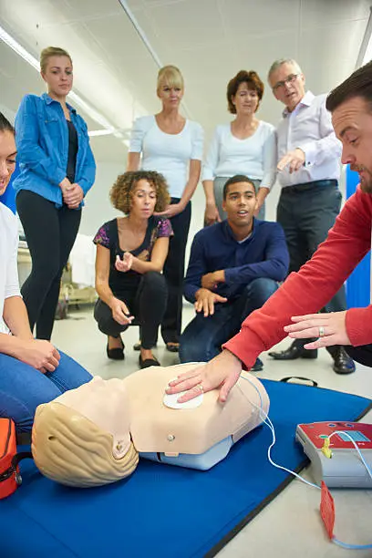a mixed age group listen to their tutor as he shows the procedure involved to resuscitate using a defibrillator .