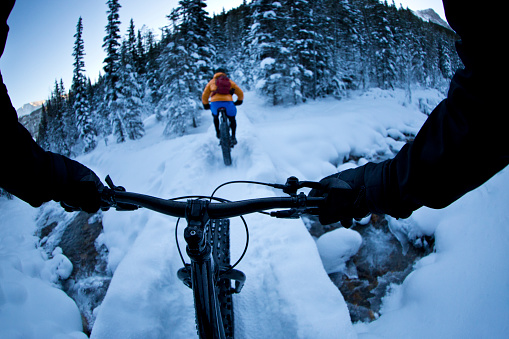 The handlebar view of two men crossing a bridge during a winter fat bike ride in the Rocky Mountains of Canada.