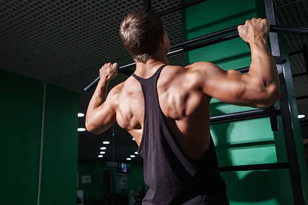 Back view of strong muscular sportsman doing pull ups in gym