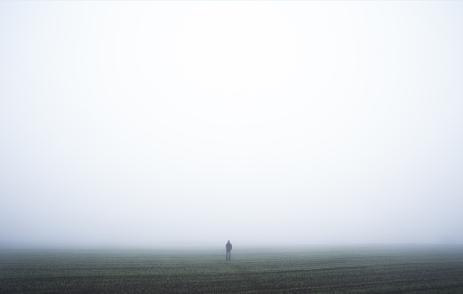 Small lonely man is standing at winter wheat field in a fog. Silhouette of man in the fog in late autumn.