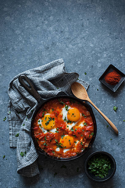 Shakshuka High resolution digital capture of shakshuka in a cast iron skillet. Shakshouka or shakshuka is a dish of eggs poached in a sauce of tomatoes, chili peppers, and onions, often spiced with cumin. In its present egg and vegetable-based form it is of Tunisian and Moroccan Jewish origin, and is now popular among many ethnic groups of the Middle East and the Maghreb. This pan of shakshuka sits on a metal surface with a grey kitchen towel, a hand carved wooden spoon, a small dish of paprika, and a small dish of parsley. moroccan culture photos stock pictures, royalty-free photos & images