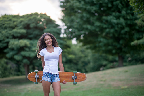 A teenage girl is walking through the park with her skateboard and is smiling while looking at the camera.