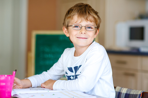 Portrait of cute happy school kid boy with glasses at home making homework. Little child writing with colorful pencils, indoors. Elementary school and education.