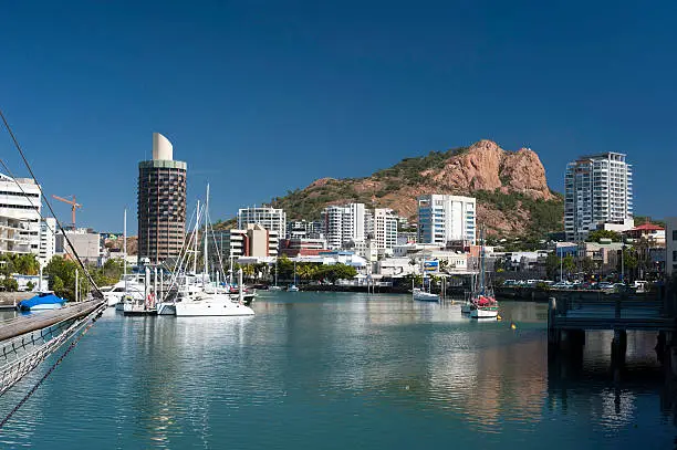 View down the quay of boats moored in the calm water of Townsville marina in Queensland, Australia with modern waterfront city central architecture and a castle hill behind