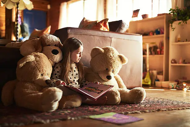 Shot of a little girl reading a book with her teddy bears around her